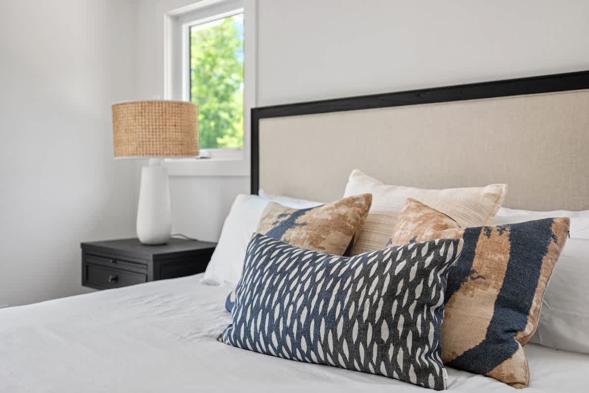 A close-up of a bed in a bedroom, featuring a neutral upholstered headboard, crisp white bedding, and an assortment of decorative pillows in blue, white, and earthy tones, with a nightstand and lamp visible.