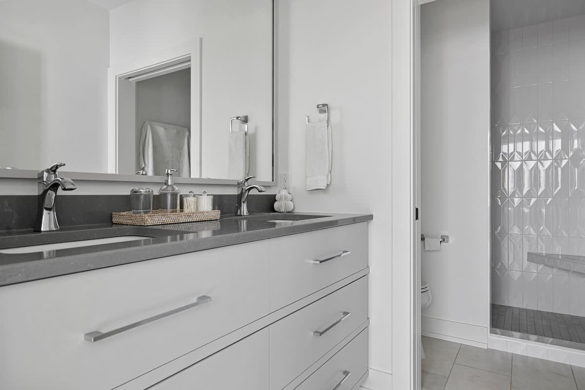 A contemporary bathroom with a double vanity featuring gray countertops, modern chrome faucets, and light gray cabinetry. A large mirror reflects the opposite wall, and a walk-in shower with white textured tiles is visible to the right.