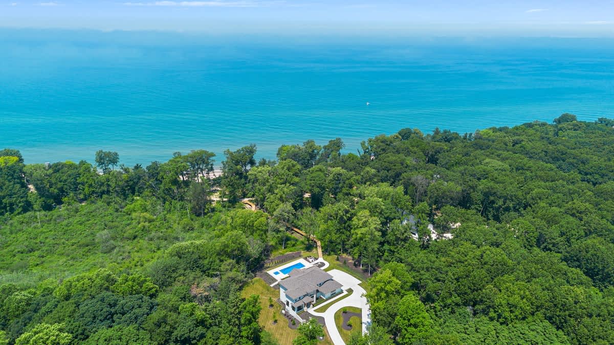 High aerial view showcasing the modern house and its pool nestled within a dense green forest, with a vast expanse of turquoise lake or ocean in the background. The property appears secluded and surrounded by nature.