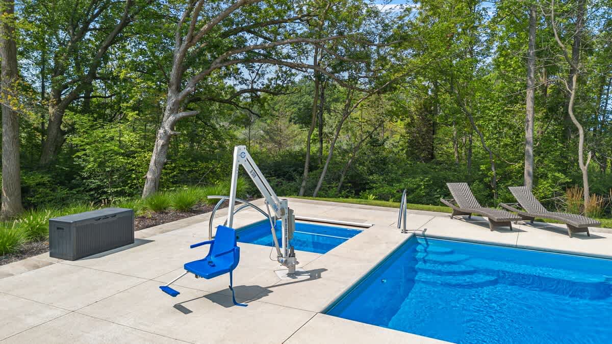 A bright blue swimming pool and an adjacent hot tub, set within a concrete patio surrounded by mature green trees. A blue pool lift for accessibility is prominently featured next to the hot tub.