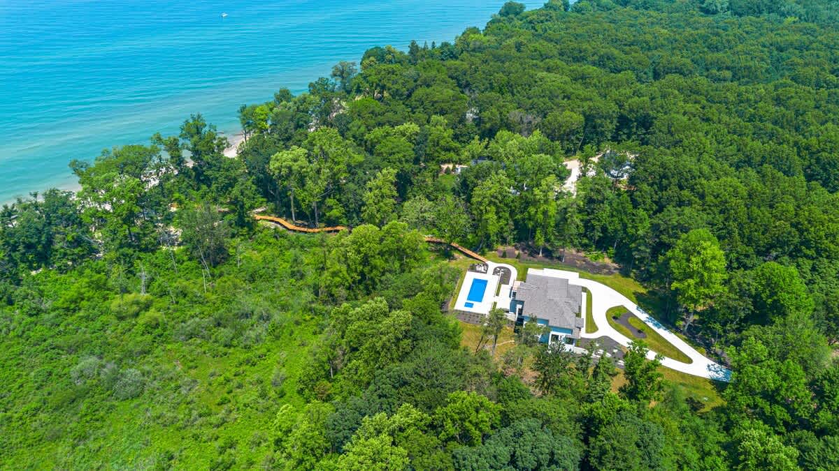 Aerial view showcasing a modern white house with a swimming pool and a long wooden boardwalk winding through dense green trees, leading towards a large body of blue water. The property is nestled within a vast forested landscape.