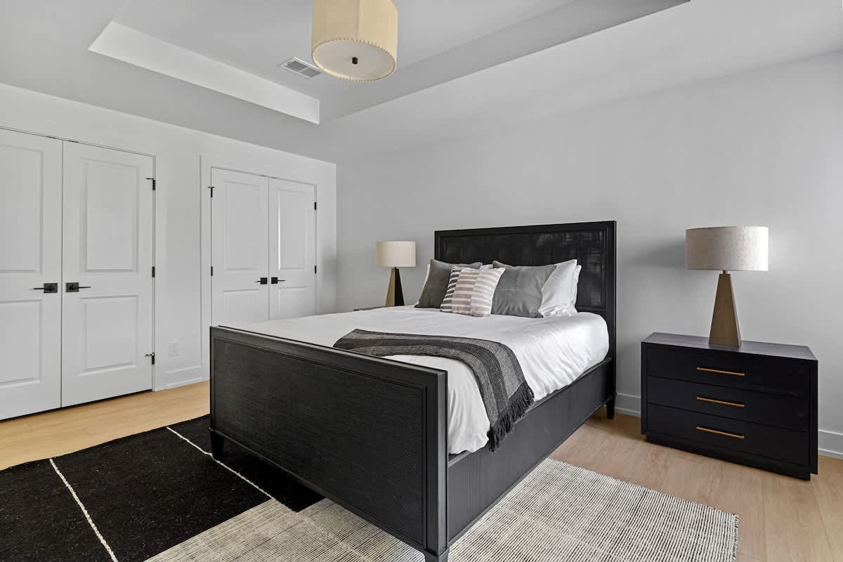 A modern bedroom featuring a black bed frame with a textured headboard, a matching black nightstand with a lamp, and a geometric black and white area rug on light wood floors. White double doors are visible in the background.