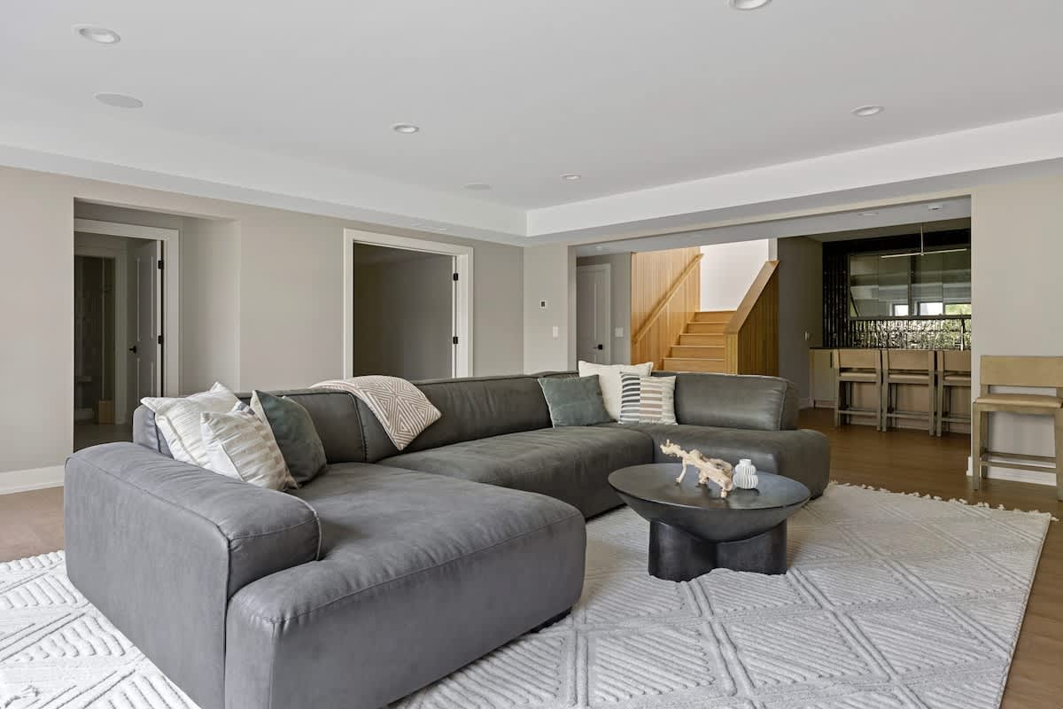 Spacious living room with light grey walls, light wood floors, and a large grey sectional sofa. A round black coffee table sits on a textured white area rug. An open doorway leads to another room, and a staircase is visible in the background.