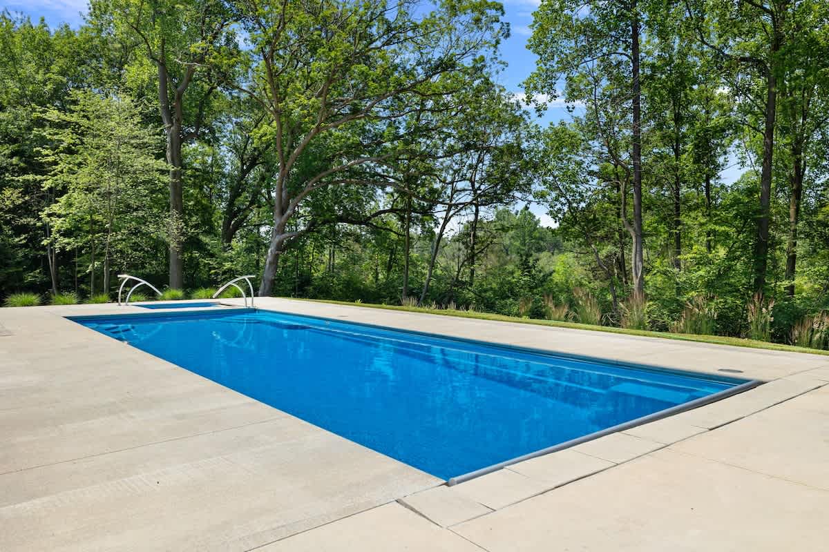 A long, rectangular bright blue swimming pool with steps at one end, surrounded by a light-colored concrete patio. The pool is set against a backdrop of dense green trees under a clear blue sky.