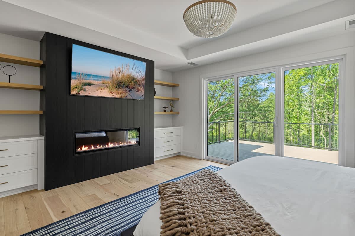 A modern bedroom featuring a bed with a brown throw, light hardwood floors, a black accent wall with a built-in fireplace and TV, floating shelves, and a large sliding glass door leading to a balcony with tree views.