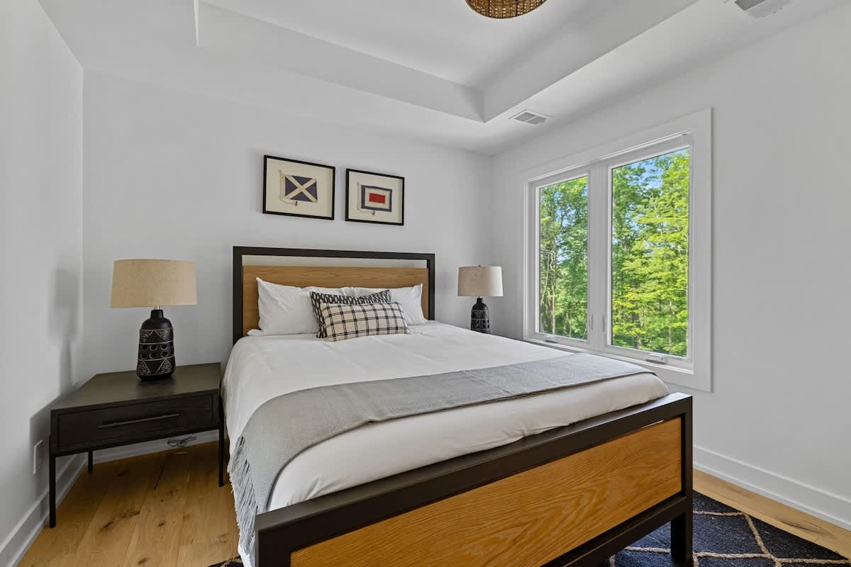 Modern bedroom with a wooden and black metal bed frame, white bedding, and a grey throw. Black nightstands with matching lamps flank the bed, and two framed nautical flag prints hang above the headboard. Large windows offer views of lush greenery.