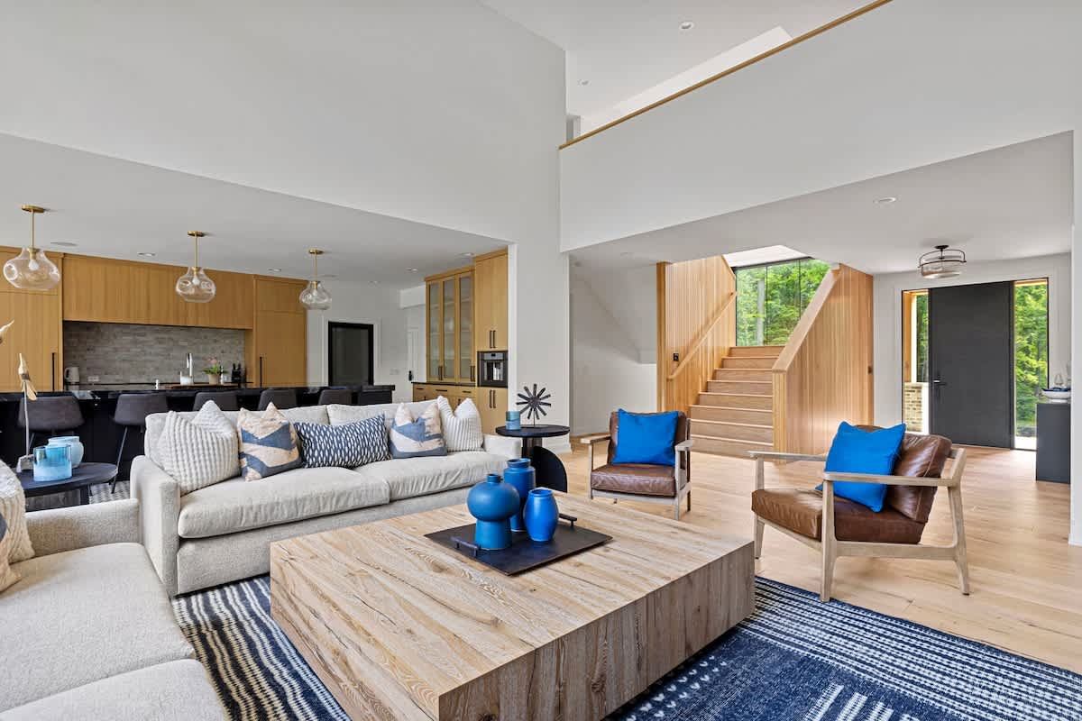 Open-concept living room with a high ceiling, light-colored sofas, brown leather armchairs, and a large wooden coffee table on a blue and white striped rug. A wooden staircase is visible in the background, leading to an upper level.