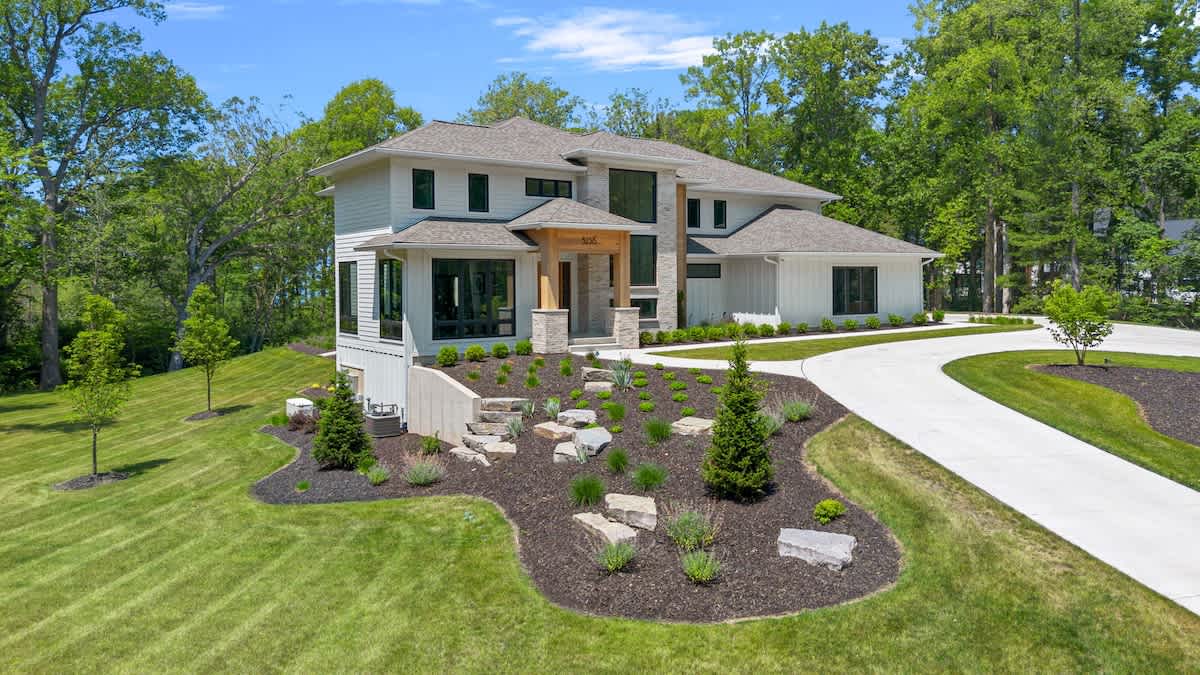 Front exterior view of a contemporary house with a light-colored facade, dark-framed windows, and a multi-level design. A winding concrete driveway leads to the entrance, surrounded by manicured landscaping with rocks and small plants.