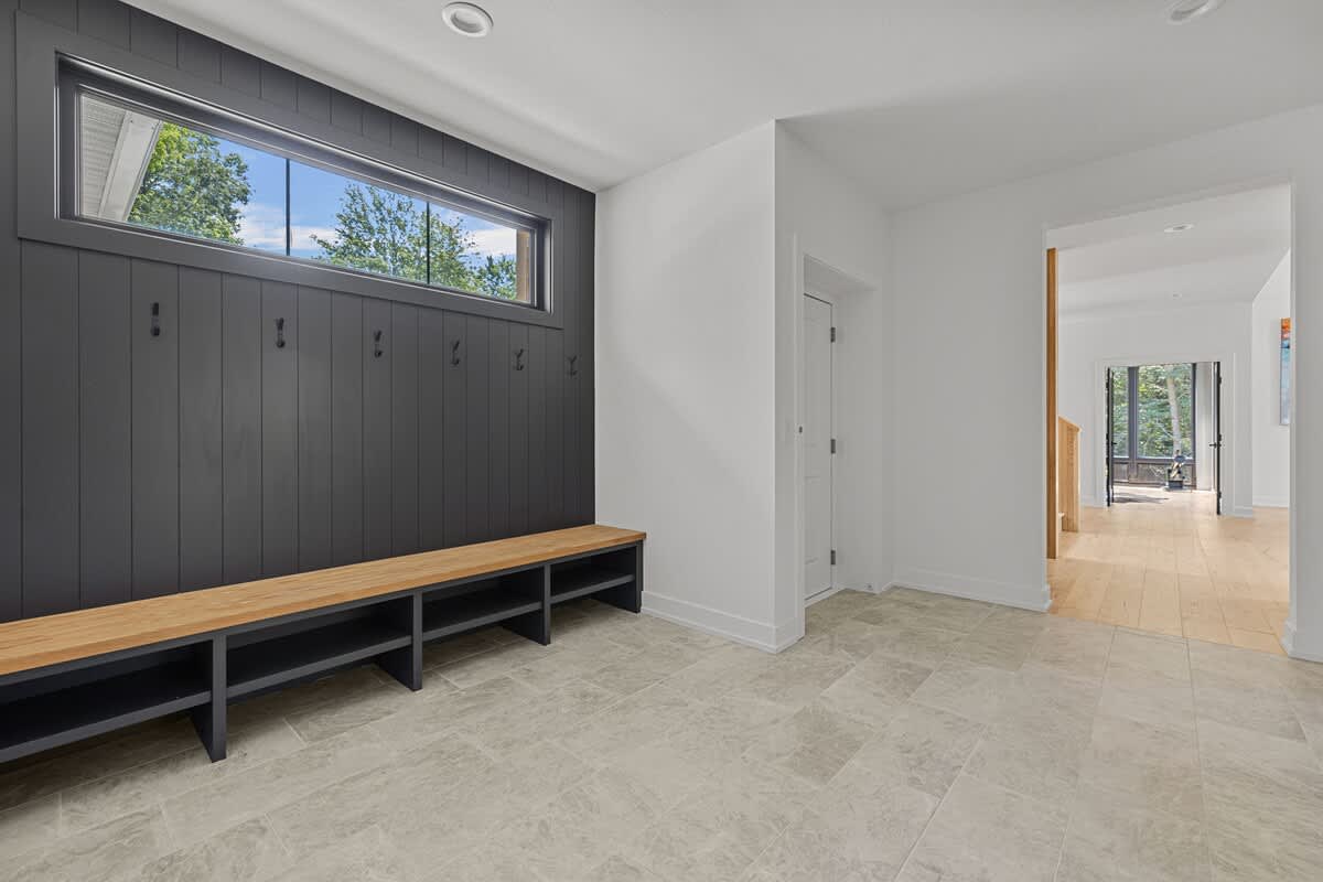 A bright mudroom with light gray tiled floors, a long wooden bench with storage cubbies, a dark gray wainscoting wall with hooks, and a horizontal window providing natural light.