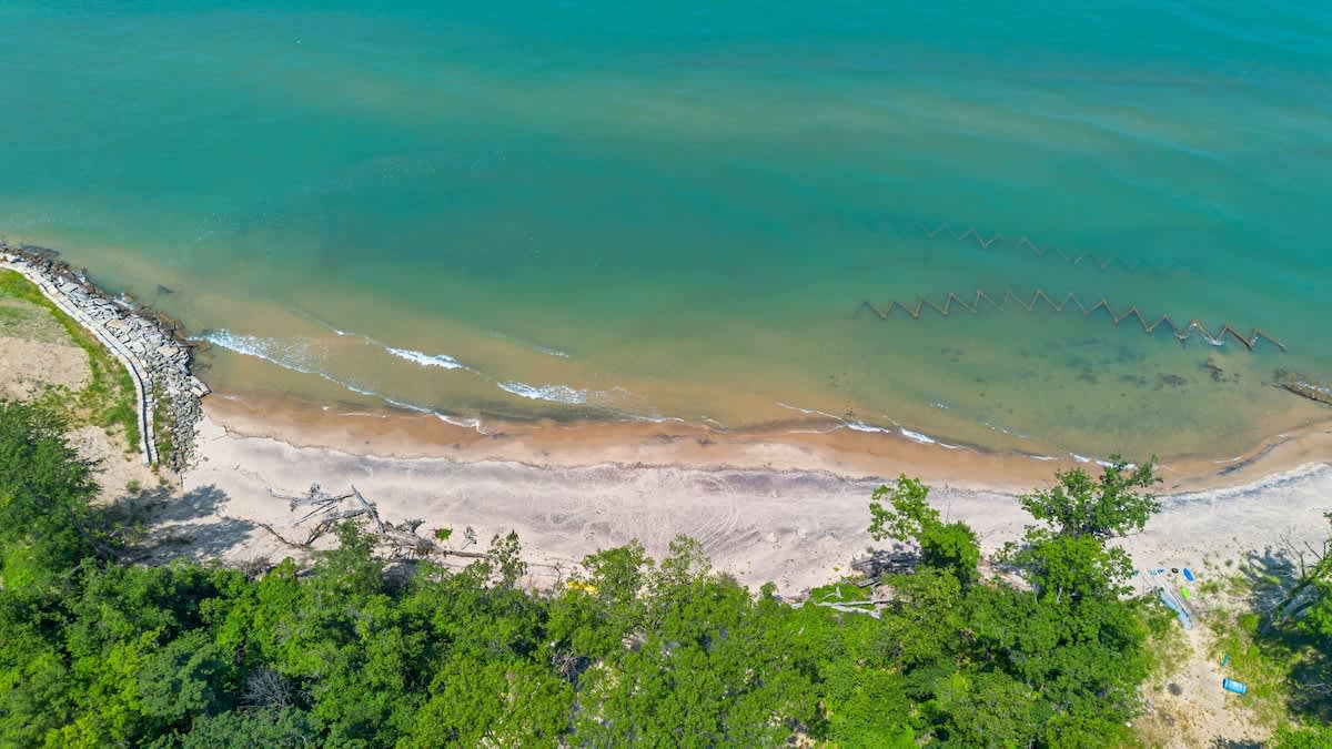 Aerial view of a private sandy beach with gentle waves meeting the clear turquoise-green lake, bordered by a lush green treeline and a rocky breakwater on the left, suggesting a serene coastal setting.