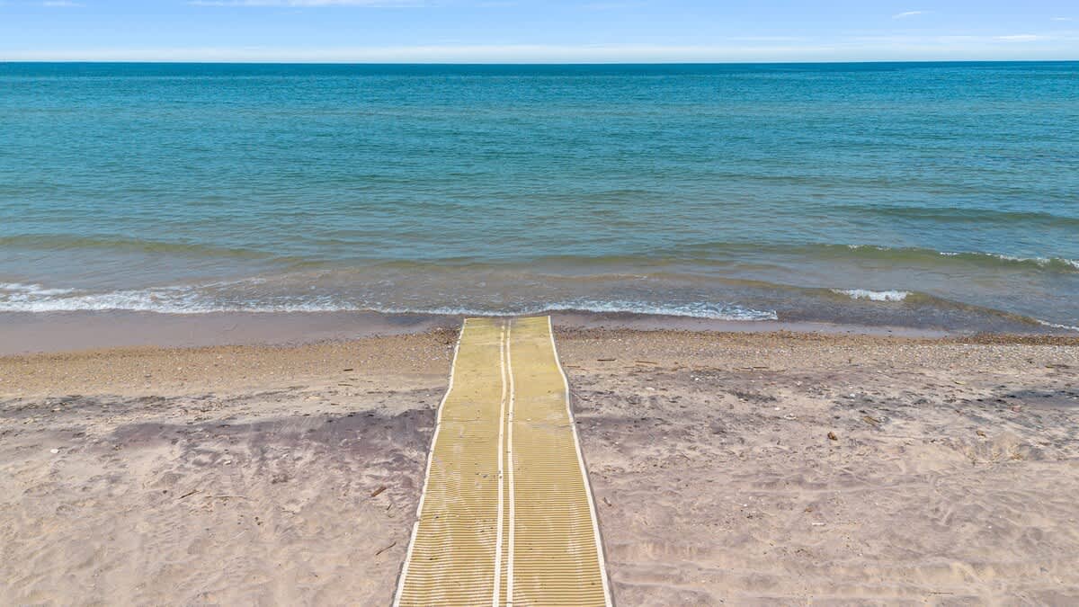 A long, yellow and white accessibility ramp extends from a sandy beach directly into the calm, blue-green waters of a lake or ocean, providing easy access to the water. The horizon shows a clear blue sky.