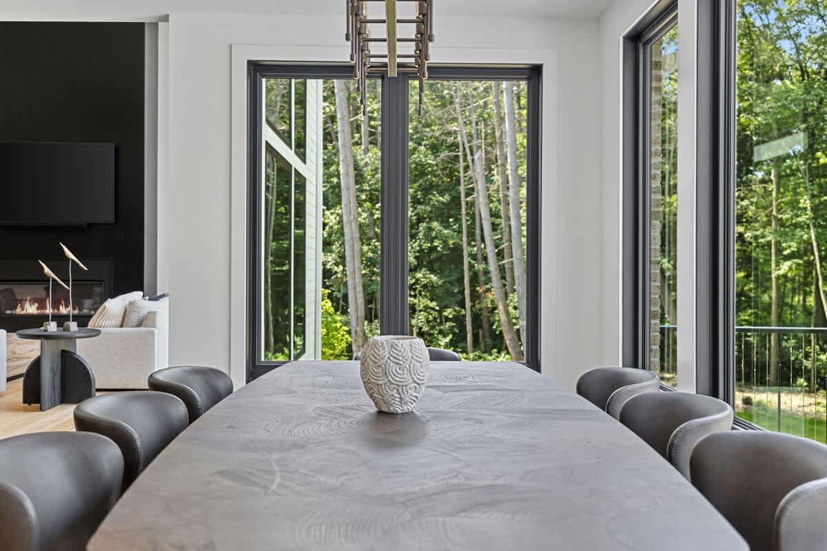 Close-up view of a long dark wooden dining table with a decorative vase, surrounded by dark chairs. Large windows provide a serene backdrop of tall trees, emphasizing the connection to nature.