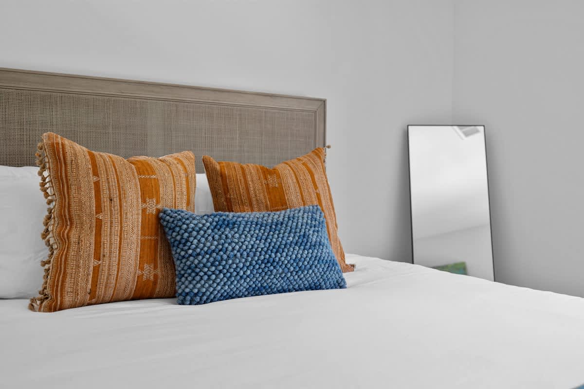 Close-up of a bed in bedroom-2 adorned with white linens, two orange textured pillows, and a blue textured lumbar pillow, with a full-length mirror in the background.