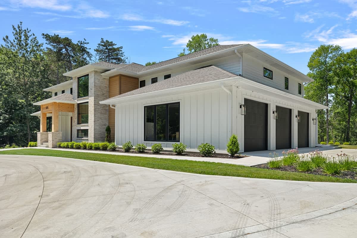Expansive front view of a modern white house with a multi-car concrete driveway, featuring a three-car garage with black doors and a mix of white siding and stone facade. Landscaped bushes line the front, with tall trees in the background.