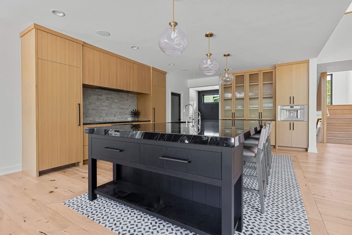 Contemporary kitchen featuring light wood cabinetry, a large black island with a dark countertop, and a patterned tile rug under the island. Pendant lights hang above, and a wooden staircase is visible in the background.