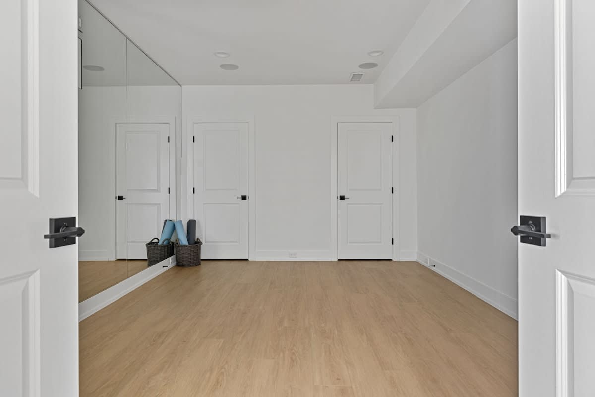 A bright, minimalist room with light wood flooring, white walls, and a large mirrored wall reflecting multiple white doors. Two woven baskets with yoga mats are visible near the mirror, suggesting a fitness or flex space.