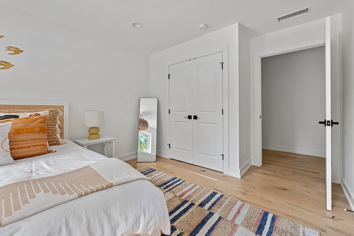 View of bedroom-3 showing a white queen bed, a colorful area rug, a white nightstand, and a closed double-door closet, with an open doorway leading to another room.
