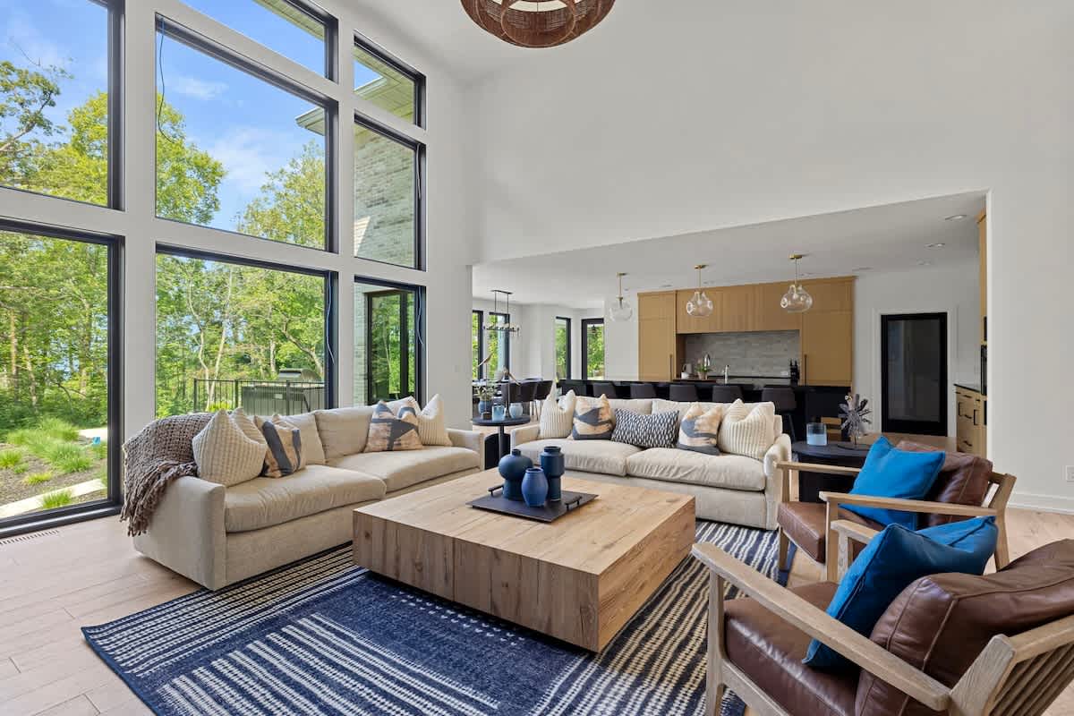 Spacious living room with a high ceiling, two light-colored sofas, two brown leather armchairs with blue pillows, and a large wooden coffee table on a blue and white striped rug. Floor-to-ceiling windows offer expansive views of trees.