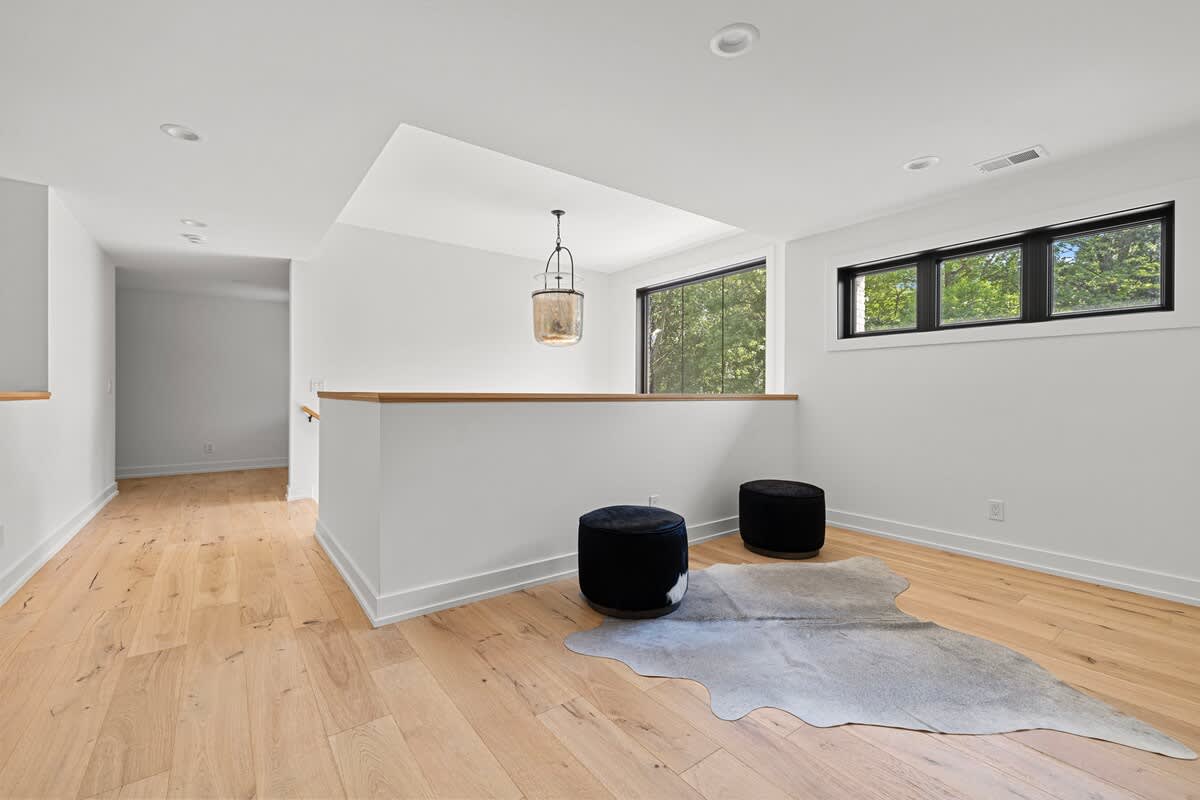 Bright open landing area with light wood floors and white walls. A low white railing separates the space, and two black velvet ottomans sit on a cowhide rug. Large windows provide natural light and views of trees.