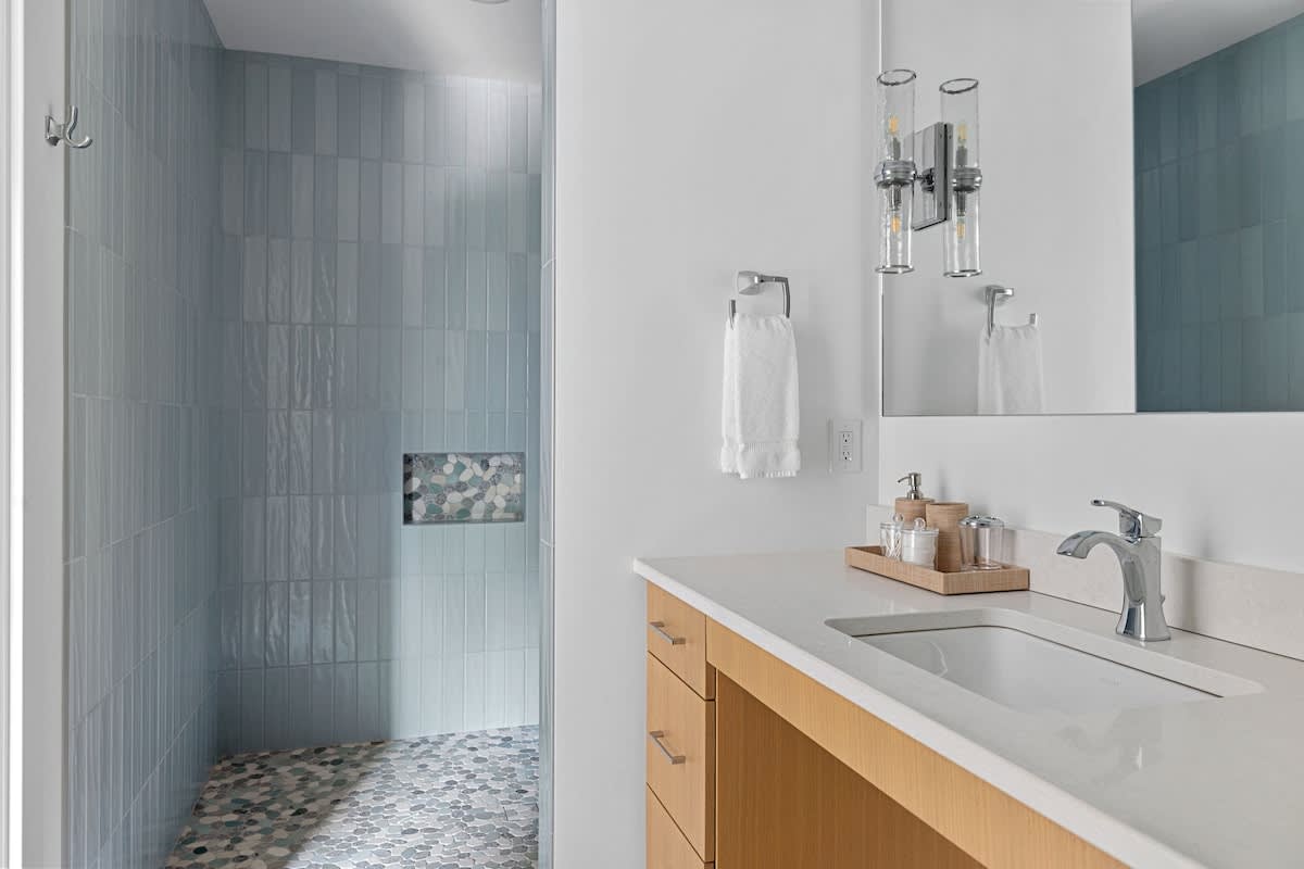 Contemporary bathroom with a light wood vanity, white countertop, and a rectangular sink. A large mirror reflects a wall-mounted light fixture. The walk-in shower features light blue vertical subway tiles and a pebble-tiled floor.