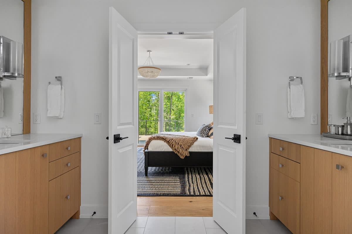 Spacious bathroom with dual light wood vanities and white countertops, looking into a bright bedroom with a large bed and outdoor views through double doors.