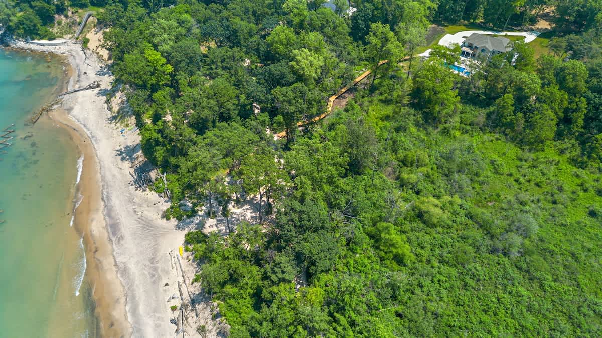 Aerial view of a sandy beach coastline with gentle waves meeting the shore, bordered by a dense green forest. A wooden boardwalk is visible winding through the trees, connecting the property to the beach.