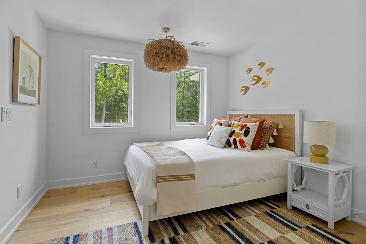 Cozy bedroom-3 with a queen bed featuring a woven headboard, a unique feathered chandelier, and two windows providing natural light and views of trees.