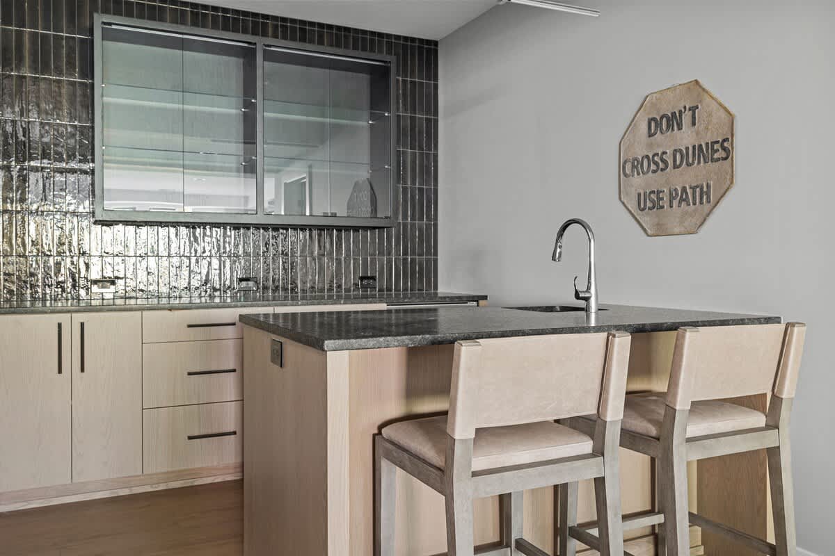 Stylish wet bar area with light wood cabinetry, dark grey countertops, and a sink with a modern faucet. The backsplash features dark, textured vertical tiles. Two bar stools are pulled up to the counter, and a 'Don't Cross Dunes Use Path' sign hangs on the wall.