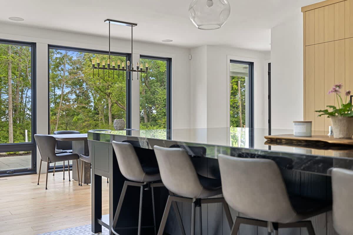 View from the kitchen island towards the dining area, featuring a long black island with bar stools. Large windows frame a lush green outdoor scene, and a modern chandelier hangs over the dining table in the distance.