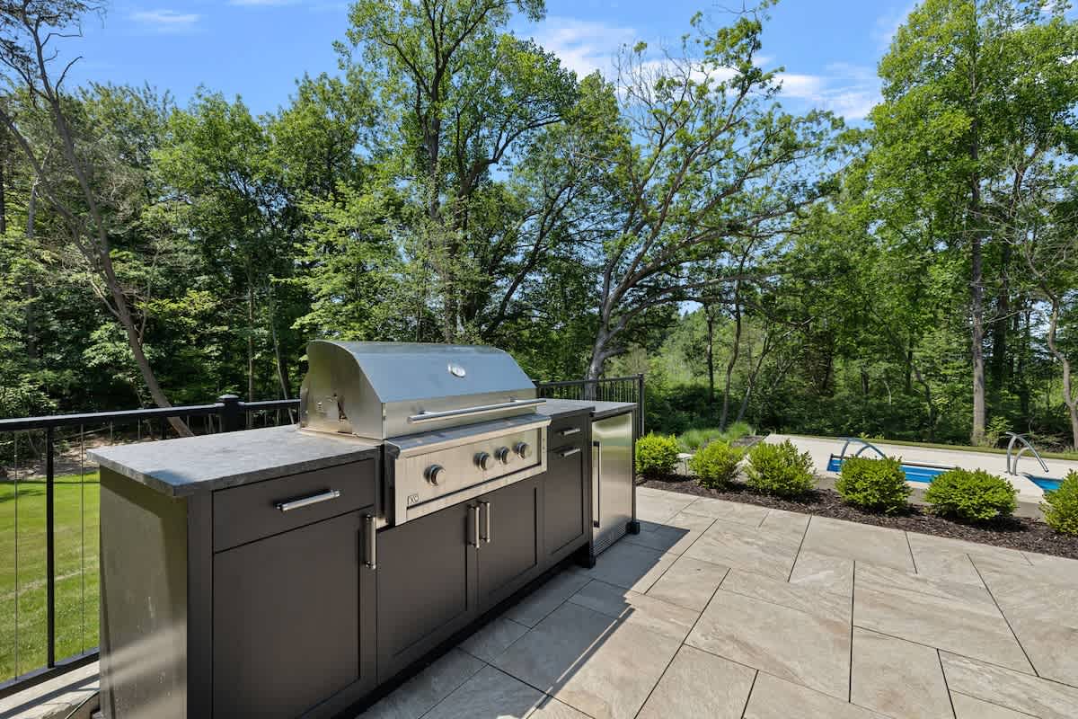 An outdoor kitchen area on a stone patio, featuring a built-in stainless steel grill and dark cabinetry with a gray countertop. Lush green bushes and trees frame the background, with a glimpse of a swimming pool.
