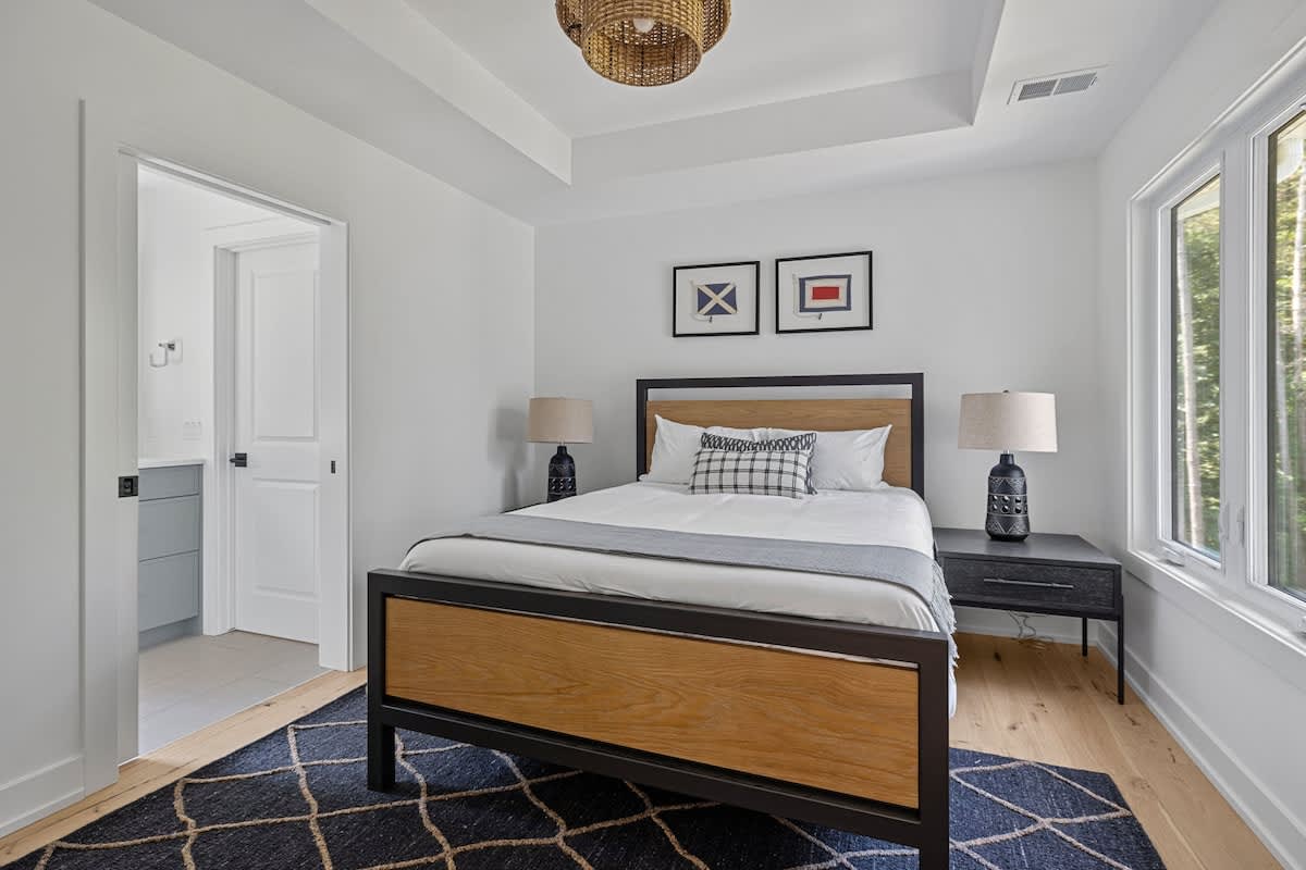 Modern bedroom featuring a wooden and black metal bed frame with white bedding and a grey throw. Two black nightstands with matching lamps flank the bed, and two framed nautical flag prints adorn the white wall. Large windows overlook green trees.