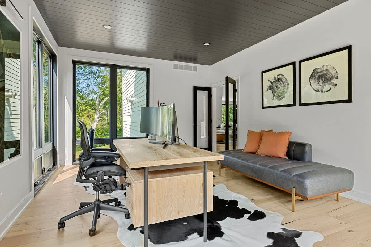Modern home office with a light wood desk, two black office chairs, a gray leather chaise lounge, and a black and white cowhide rug on light wood floors. Large windows offer views of greenery, and the ceiling is dark gray.