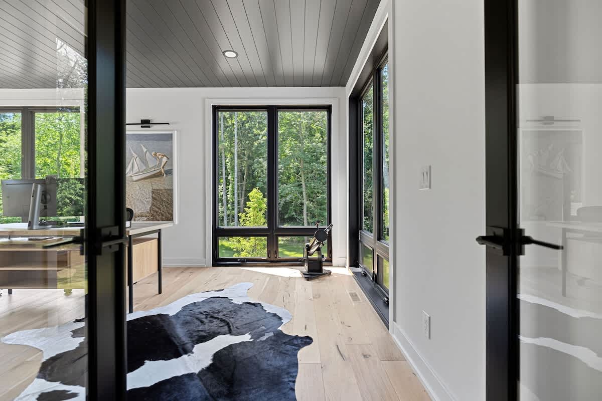 Modern office space with light hardwood floors, a dark paneled ceiling, and large black-framed windows offering views of lush greenery. The room features a desk, a cowhide rug, and glass-paned double doors.
