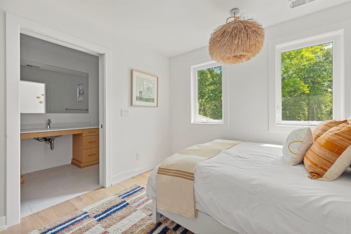 Bright bedroom with white walls, light wood floors, and a bed with white and orange bedding. A unique straw-like pendant light hangs from the ceiling, and two large windows offer views of lush greenery. An open doorway leads to a bathroom with a vanity.