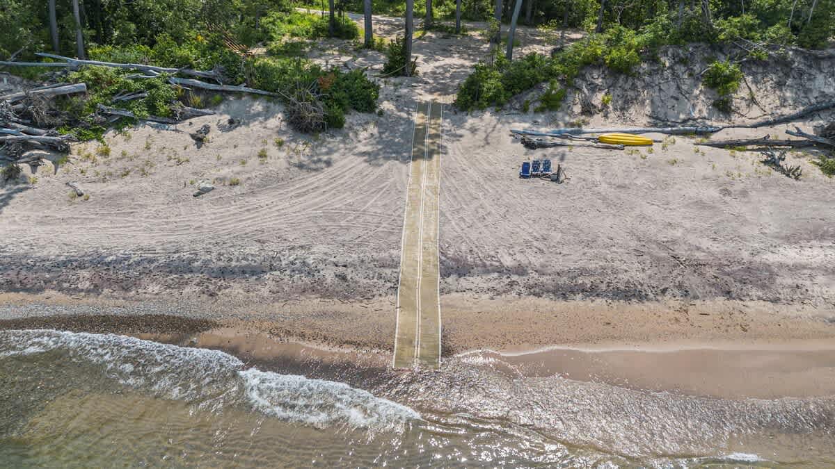 Aerial view of a sandy beach with a yellow and white accessibility ramp leading into the water, flanked by two yellow kayaks and beach chairs. The beach is surrounded by green trees and shows tire tracks in the sand.