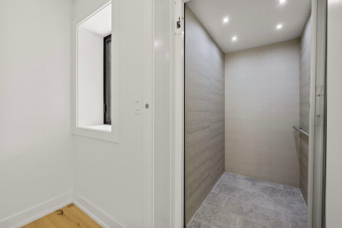 An interior view of a modern elevator with textured gray walls and recessed lighting, situated in a hallway with light hardwood floors and a small window.