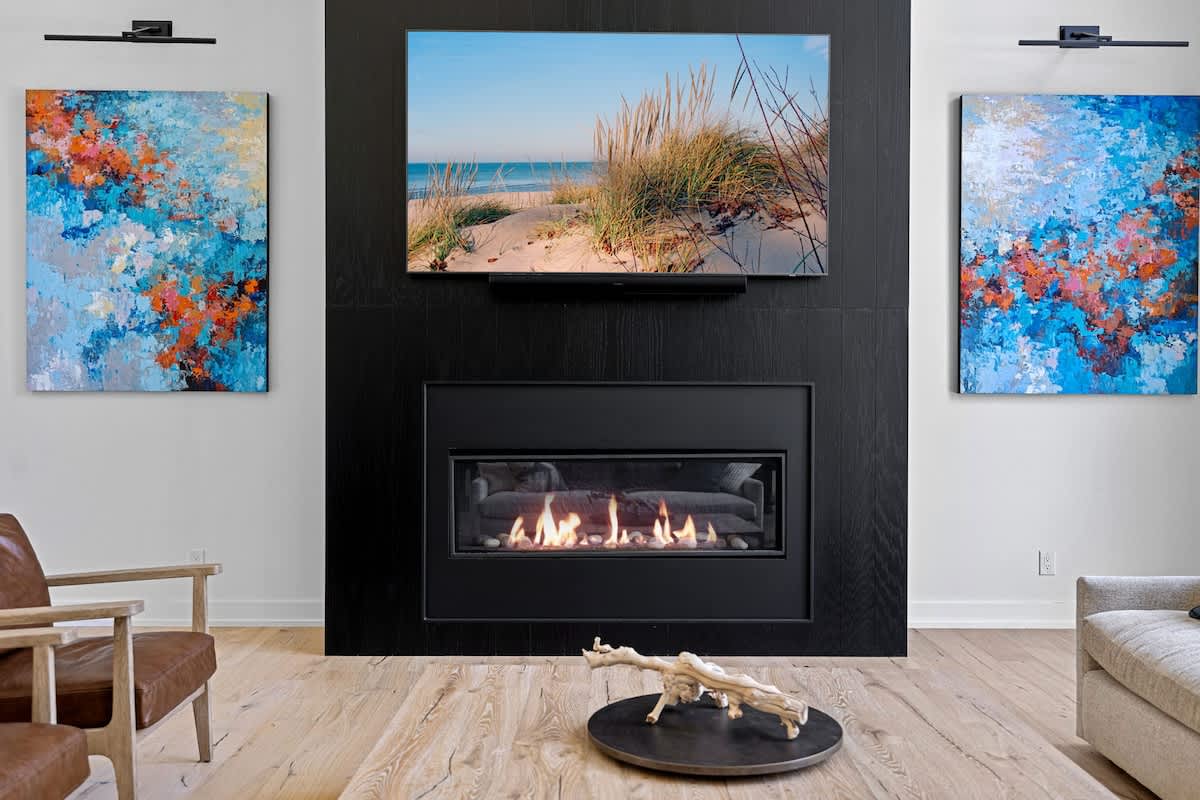 Contemporary living room featuring a black fireplace with a mounted TV above it, flanked by two abstract paintings with blue and orange tones. Light wood floors and a dark circular coffee table with decorative wood complete the space.