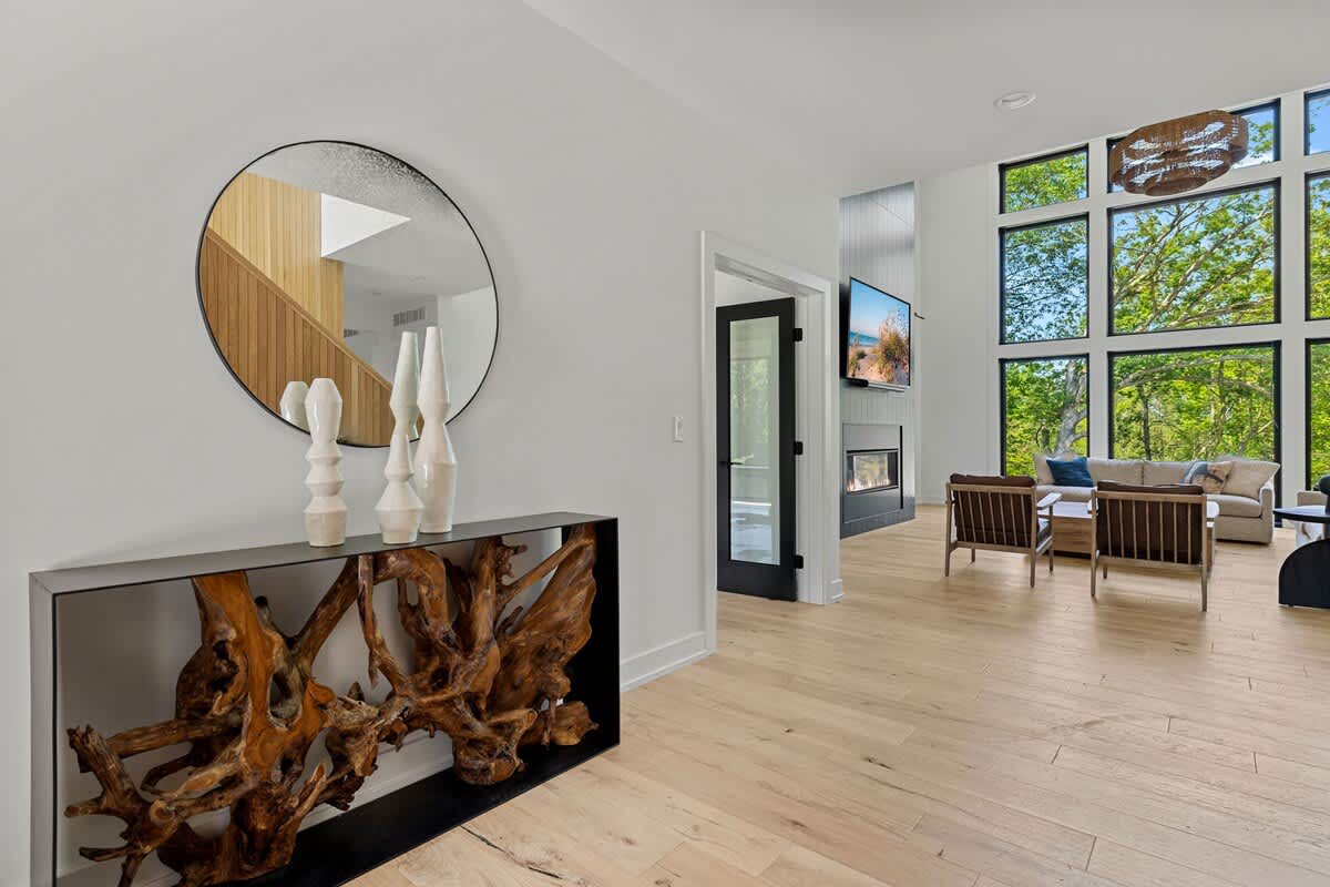 View from the foyer into the living room, highlighting a console table with a large round mirror and sculptural decor. Light hardwood floors extend throughout, with a glimpse of the living room's tall windows and fireplace.