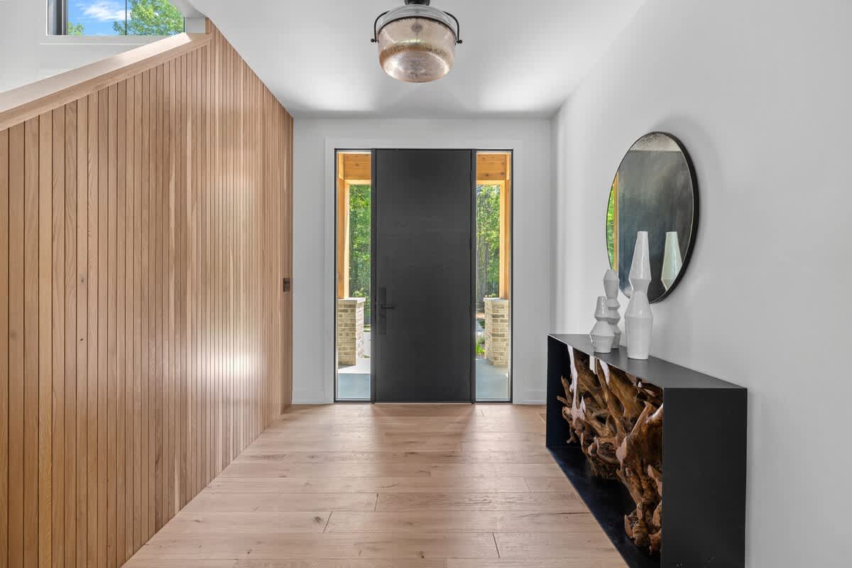 Bright foyer with light hardwood floors and a tall wooden slatted wall leading to a staircase. A modern black front door with side windows is visible, along with a console table featuring a large round mirror and decorative vases.