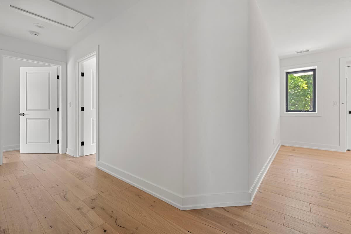 A clean, minimalist hallway with light hardwood floors, white walls, and white doors, featuring a small window that provides natural light to the space.
