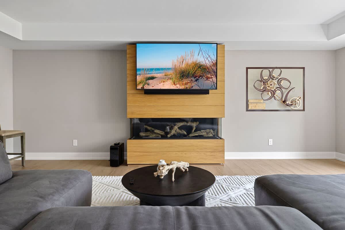 Spacious media room with light grey walls and light wood floors. A large flat-screen TV is mounted above a modern linear fireplace, both integrated into a light wood paneling feature wall. Grey sectional sofas and a round coffee table furnish the space.