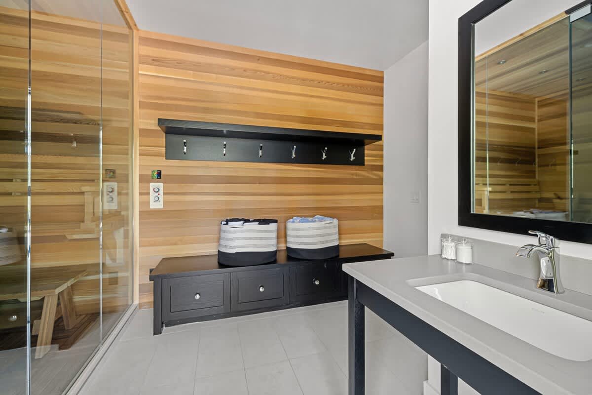 A spa-like room featuring a wooden sauna with glass walls, a vanity with a light gray countertop and a black framed mirror, and a black storage bench with hooks on a wood-paneled wall.