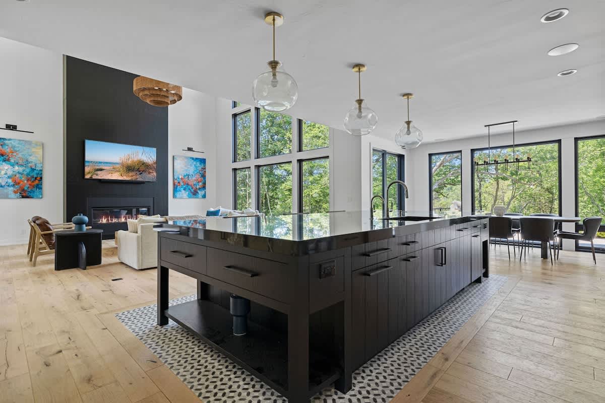 Expansive open-concept living area with a black kitchen island, light wood floors, and a black fireplace accent wall. Large windows overlook lush greenery, connecting the living room, kitchen, and dining areas.