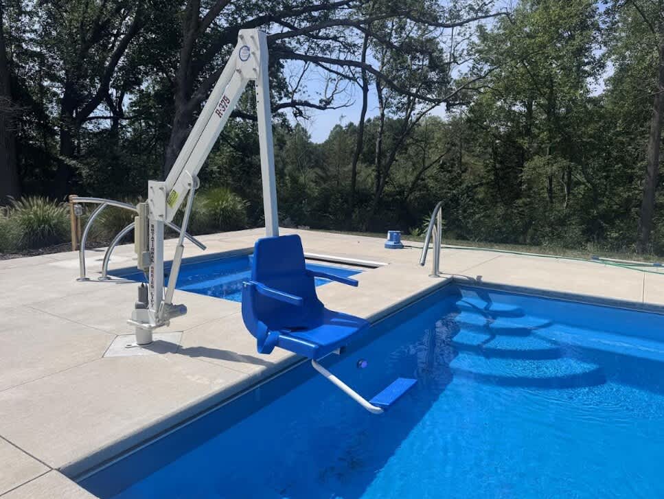 A blue pool accessibility lift with a seat is positioned over the edge of a bright blue swimming pool, set against a backdrop of green trees and a clear sky. The concrete pool deck is visible around the pool.