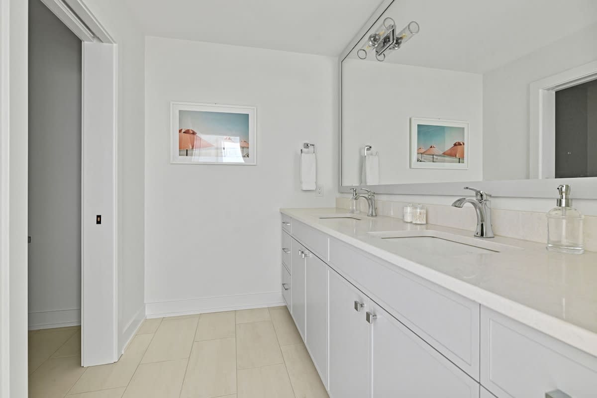 Bright bathroom featuring a long white double vanity with light grey cabinets and two rectangular sinks. A large mirror spans the length of the vanity, reflecting wall-mounted light fixtures. White tiled floors complete the clean, modern look.