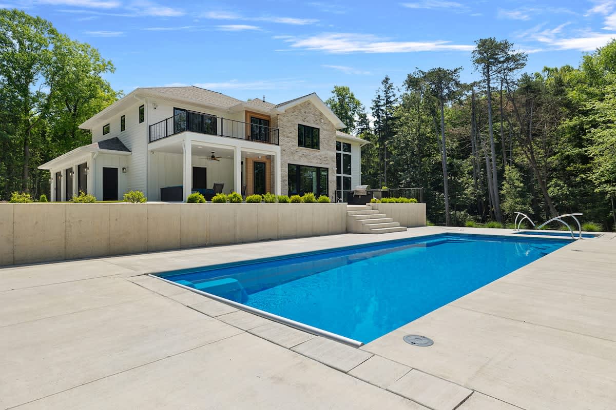 Modern white house with black accents and stone facade, featuring a large rectangular swimming pool with concrete decking, surrounded by lush green trees under a bright blue sky. A raised patio area with outdoor seating is visible behind the pool.