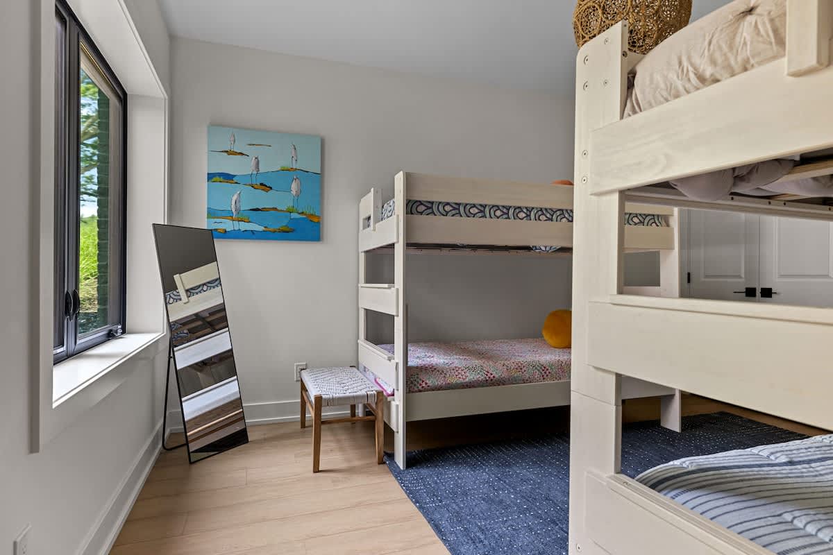 A children's bedroom featuring light wood bunk beds with blue and white bedding, a blue area rug, and a full-length mirror leaning against the wall. A colorful abstract painting hangs above a small stool.