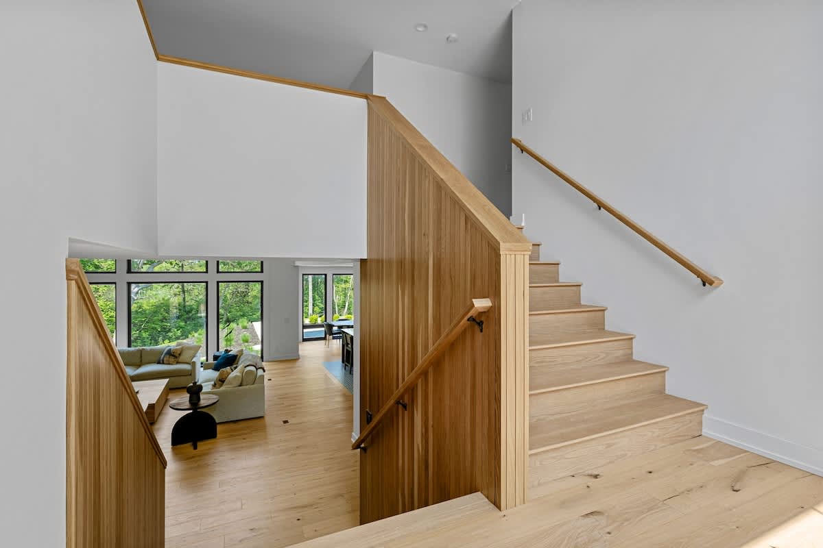 A modern entryway featuring a light wooden staircase with matching railings, leading down to a spacious living area with large windows overlooking a lush green landscape.