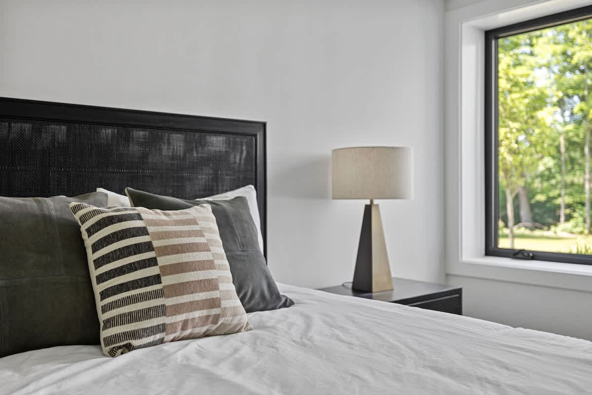 A close-up of a modern bed with a black textured headboard, white bedding, and decorative pillows, next to a black nightstand with a beige lamp. A large window with a black frame offers a view of lush greenery outside.