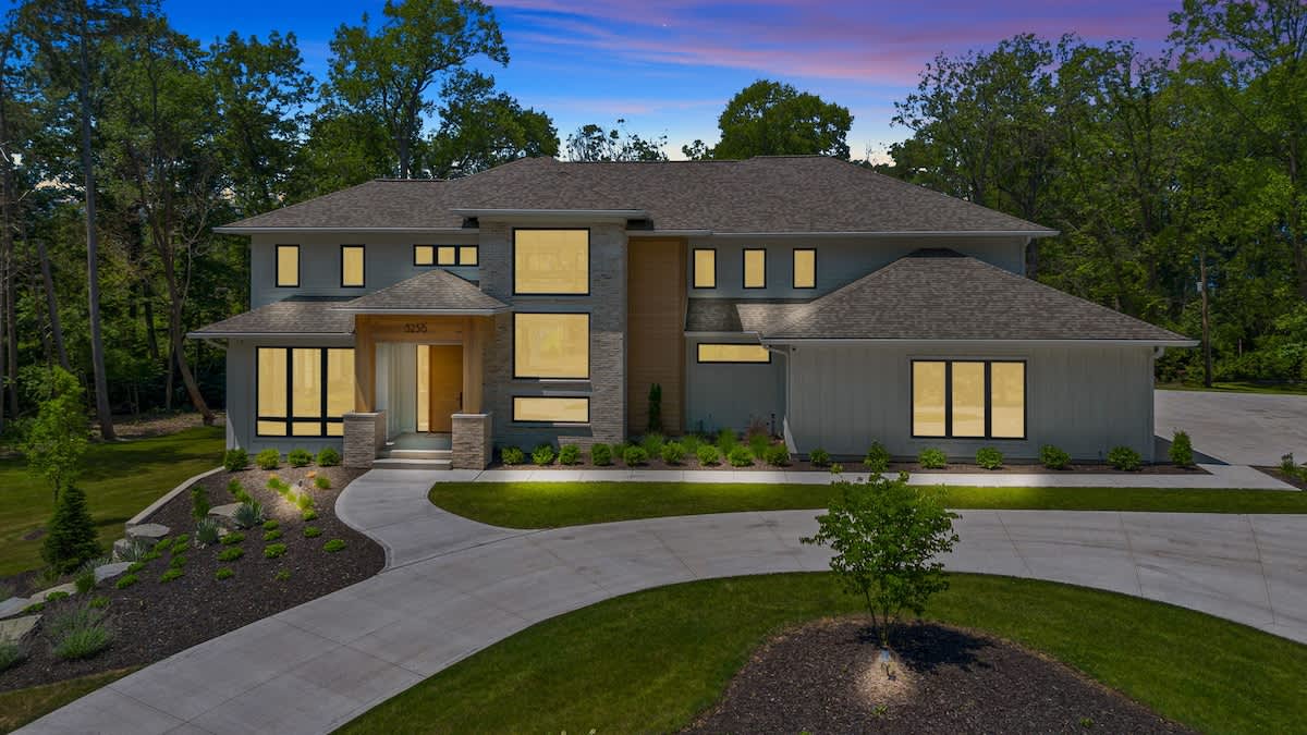 Front exterior view of a contemporary house at dusk, showcasing warm illuminated interior spaces through large windows, a stone and wood facade, and a circular driveway surrounded by lush landscaping and mature trees.